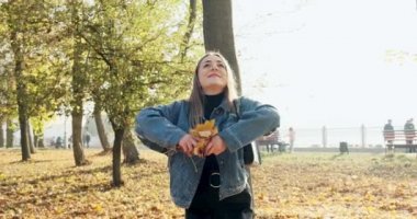 Mutlu genç bir kadın güneşli sonbahar parkında sarı yapraklar fırlatırken eğleniyor. Yavaş çekim