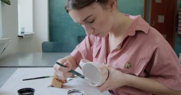 Γυναίκα αγγειοπλάστη κάθεται και ανακατεύει χρώμα με ένα πινέλο ένα φλιτζάνι στο τραπέζι. Γυναίκα που φτιάχνει κεραμικά αντικείμενα. Εργασίες αγγειοπλαστικής, χειροποίητες και δημιουργικές δεξιότητες — Αρχείο Βίντεο
