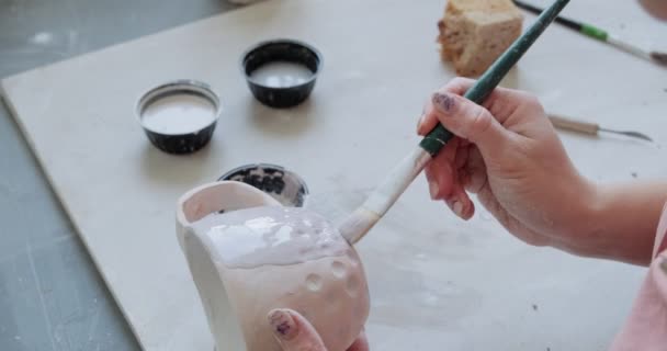 Female potter sitting and stirs paint with a brush a cup on the table. Woman making ceramic item. Pottery working, handmade and creative skills — Stock Video