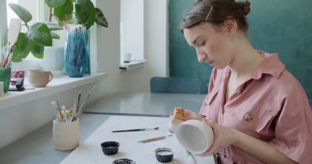 Ceramista femminile seduta e mescola dipingere con un pennello una tazza sul tavolo. Donna che fa oggetti in ceramica. Lavorazione della ceramica, abilità artigianali e creative — Video Stock