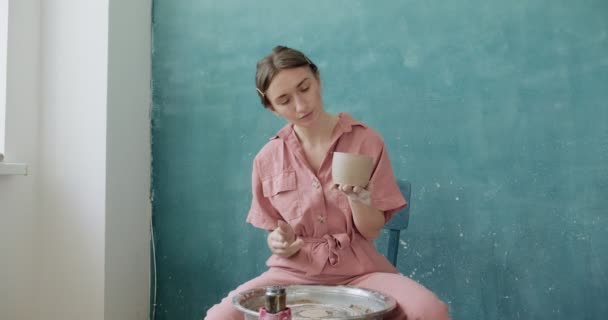 Γυναίκα αγγειοπλάστη κάθεται και κάνει ένα φλιτζάνι στον κεραμικό τροχό. Γυναίκα που φτιάχνει κεραμικά αντικείμενα. Εργασίες αγγειοπλαστικής, χειροποίητες και δημιουργικές δεξιότητες — Αρχείο Βίντεο