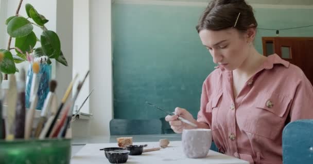 Ceramista femminile seduta e mescola dipingere con un pennello una tazza sul tavolo. Donna che fa oggetti in ceramica. Lavorazione della ceramica, abilità artigianali e creative — Video Stock