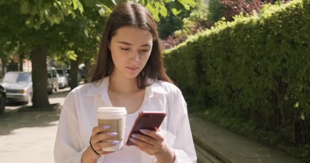 Mujer joven con llevar la taza y beber café caliente, mirando el teléfono inteligente, vestido con traje de negocios cerca de la pared del arbusto — Vídeo de stock