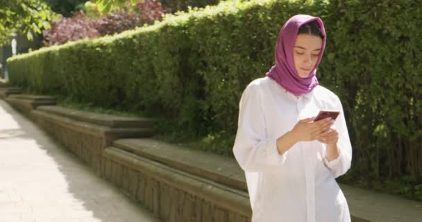 Hermosa mujer joven mirando el teléfono inteligente, usando el pañuelo de cabeza tradicional. Atractiva hembra en hijab — Vídeos de Stock