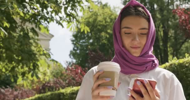 Hermosa mujer joven beber café y mirando el teléfono inteligente, usando el pañuelo de cabeza tradicional. Atractiva hembra en hijab — Vídeos de Stock