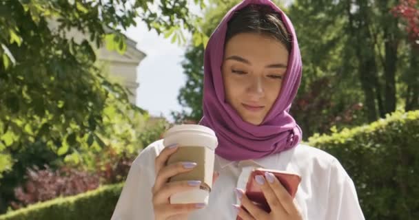 Hermosa mujer joven beber café y mirando el teléfono inteligente, usando el pañuelo de cabeza tradicional. Atractiva hembra en hijab — Vídeos de Stock