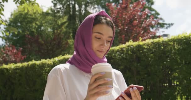 美しい若い女性は、伝統的なスカーフを着て、コーヒーを飲み、スマートフォンを見ています。ヒジャーブの魅力的な女性 — ストック動画