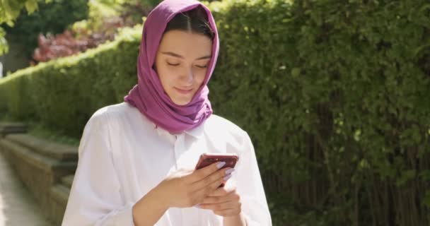 Hermosa mujer joven mirando el teléfono inteligente, usando el pañuelo de cabeza tradicional. Atractiva hembra en hijab — Vídeos de Stock