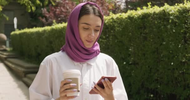 Caminar hermosa mujer joven beber café y mirando el teléfono inteligente, usando el pañuelo de cabeza tradicional. Atractiva hembra en hijab — Vídeos de Stock
