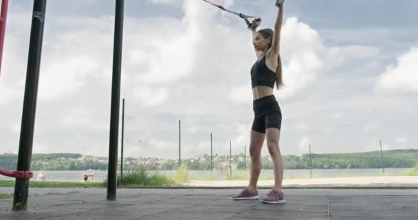 Hermosa mujer joven hace el entrenamiento con correas de fitness al aire libre. Atractivo entrenamiento femenino. TRX — Vídeo de stock