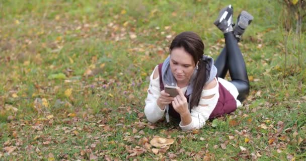 Счастливая девушка лежит в парке на траве. Фотографии по телефону. Желтая листва — стоковое видео