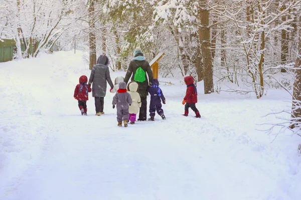 Grupo de niños pequeños irreconocibles con maestros de jardín de infantes, de vuelta a nosotros, caminar tomados de la mano en el parque cubierto de nieve, ocio de invierno, estilos de vida activos — Foto de Stock
