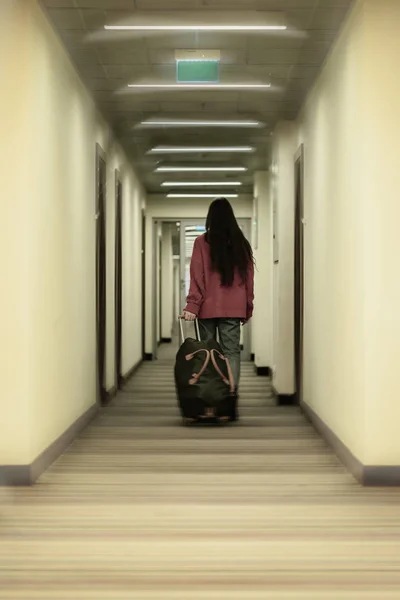 Unrecognizable girl back to us is on the corridor of the hotel holding a travel bag in her hand