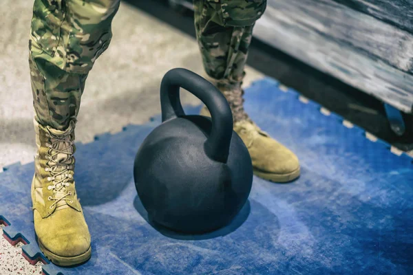 Legs of abstract athlete in camouflage pants and in yellow boots near kettlebell closeup, preparation before heavy lifting weight