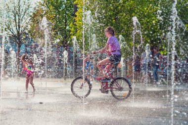Yaz, sıcak, çeşmeler arasında bisiklete binen bir kız, insanlar su jetleri serin