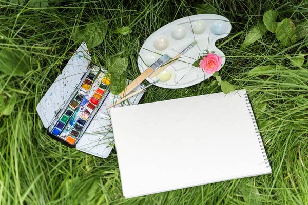 Schetsboek, aquarelverf en het kunstenaarspalet liggen in het gras, bovenaanzicht — Stockfoto