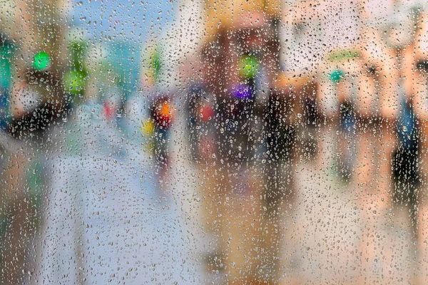 Blurred motion abstract background. Raindrops on window glass. Cloudy evening in a modern city, people walk in the street, view through window with droplets. Concept of rainy weather, seasons, modern lifestile.