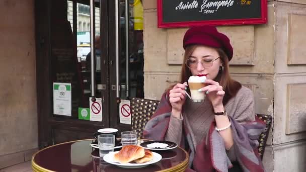 美丽的女孩长头发 红色的口红 贝雷帽坐在法国咖啡馆吃早餐 喝咖啡与羊角面包和微笑 — 图库视频影像
