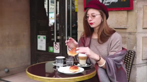 长头发 红口红 贝雷帽在法国咖啡馆吃早餐 喝咖啡和羊角面包 微笑的年轻女孩 — 图库视频影像