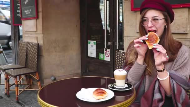 漂亮的女孩长头发 红色的口红 贝雷帽坐在法国咖啡馆吃早餐 喝咖啡与羊角面包和微笑 — 图库视频影像