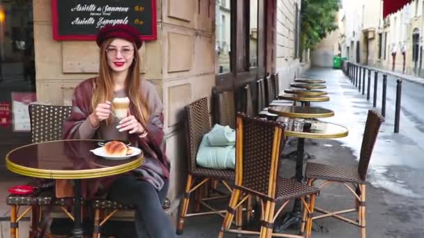 漂亮的女孩长头发 红色的口红 贝雷帽坐在法国咖啡馆吃早餐 喝咖啡与羊角面包和微笑 — 图库视频影像