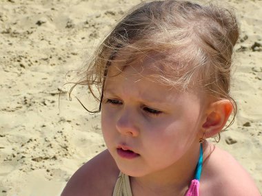 Kumlu bir plajın arka planında üzgün üzgün bakan küçük kız.