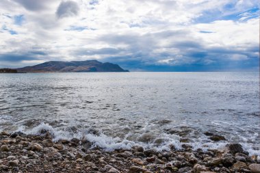Küçük dalgalar sert çakıllı deniz kıyısına çarpıyor. Uzakta, Cape Meganom. Kırım