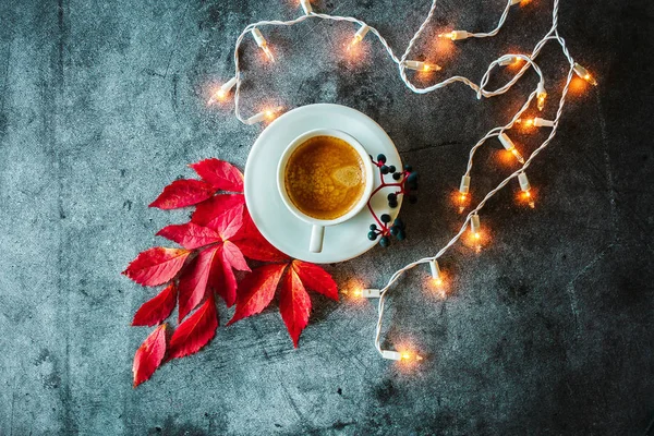 Cup of coffee, dark berries and red autumn leaves on a gray dark background. Photo above. Autumn still life. Object shooting. Leaf pattern. Decoration with a luminous garland