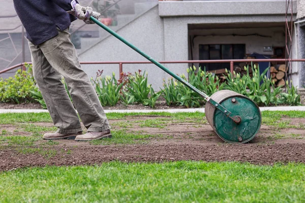 L'uomo in divisa da lavoro tira rullo prato dietro. Necessità dopo lungo inverno e primavera per superficie piana. Paesaggio sul giardino. Lavoratore estivo . — Foto Stock