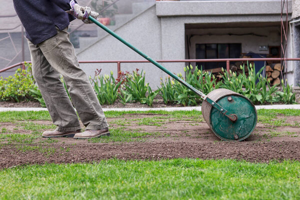 Man in working outfit pulls lawn roller behind. Necessity after long winter and spring for flat surface. Landscaping on the garden. Summer worker. 
