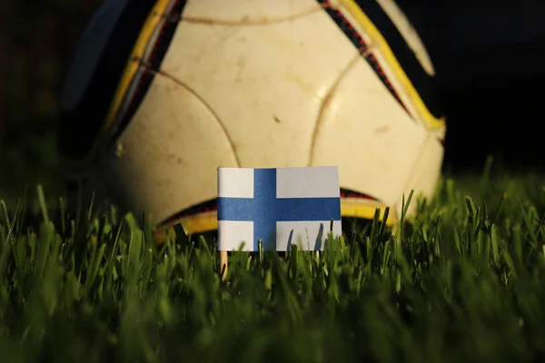 Finland flag stick to grass with football ball in background. Soccer championship 2022. Euro 2020. Blue and white paper on wooden stick. Finnish state symbol. International action