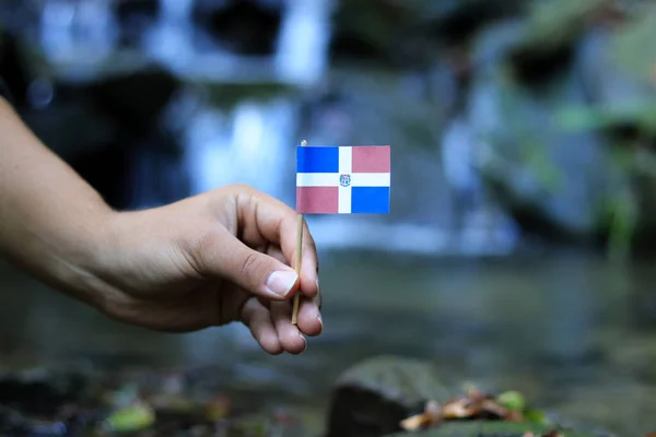 Bandera de República Dominicana en ambiente acuático. Mano del joven sostiene este símbolo estatal cerca de la cascada y el río. Concepto de humanidad y naturaleza. Protección de la naturaleza —  Fotos de Stock