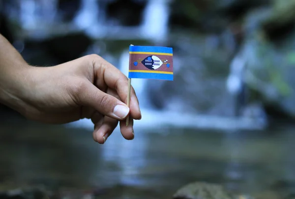 One's hand holds and waves with national flag of Eswatini on wooden stick. State symbol in water and wet environment. Prove of humanity and maturity. Concept of abundance and beauty of nature — Stock Photo, Image