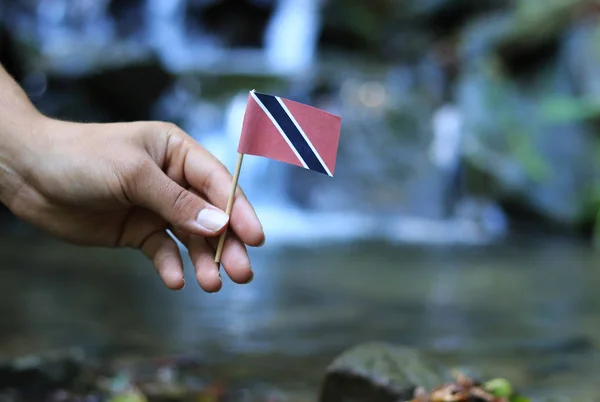 Poor land Trinidad and Tobago in the hands of affluent people. Young man holds flag of Trinidad and Tobago near stream. Concept of humanity and dominance. Prove of depraved and avariciousness — Stock Photo, Image