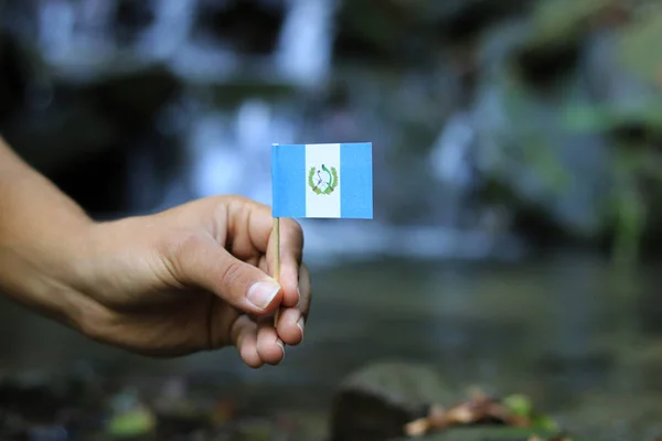 Bandiera nazionale blu e bianca del Guatemala in persona. Colore uomo tiene bandiera su bastone di legno e onde. Acqua e cascate sullo sfondo. Concetto di umanità e onniscienza — Foto Stock