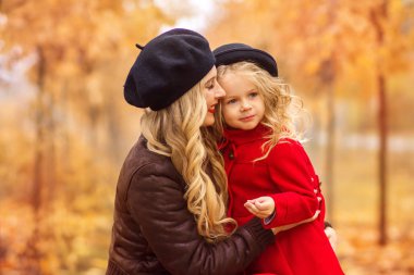 siyah bere ve kırmızı bir uzun elbise güzel bir anne yavaşça kulaklı küçük bir kızı parlak kırmızı ceket ve siyah şapka bir altın sonbahar bahçe arka planda hugs..