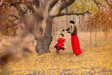 küçük kızı menteşeli bir salıncak üzerinde kırmızı paltolu bir eski gnarly ağacının sonbahar bir bahçede kırmızı elbiseli Genç bir anne sallıyor.