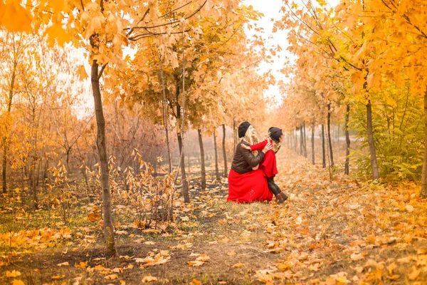 黒ベレー帽と赤のロングドレスで美しい母は優しくゴールデン秋の庭の背景に耳を持つ明るく赤いコートと黒の帽子の小さな娘を抱擁します — ストック写真