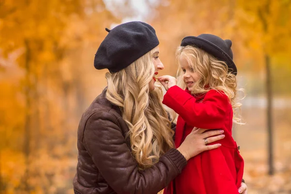 一个美丽的母亲在黑色贝雷帽和红色长裙轻轻拥抱一个小女儿在明亮的红色大衣和黑色的帽子 在金色的秋季花园的背景下的耳朵 — 图库照片