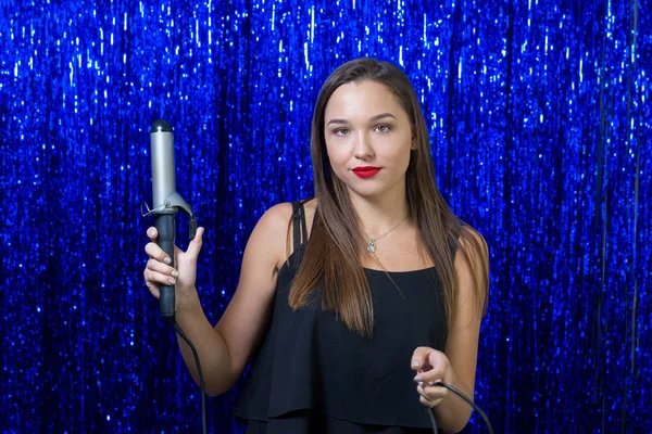 sexy girl stylist with red lipstick on her lips is holding a professional hair curling iron for modeling hair and posing positively on the camera on a background of shiny blue sequins.