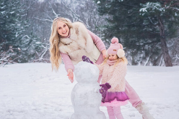 mom blonde and little daughter dressed in pink clothes have fun and make a snowman in a snowy park