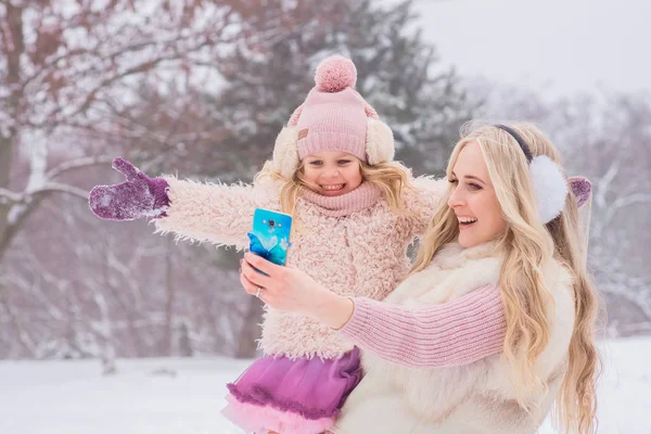 pembe elbise giymiş anne Sarışın ve küçük kızı eğlenmek ve karlı bir parkta bir cep telefonunda selfie yapmak
