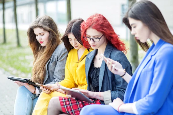 Quattro ragazze con gadget sono sedute sulla panchina. Il concetto di Internet, social network, studio e stile di vita — Foto Stock