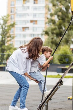 Kadın, genç oğlunu modern bir şehir oyun parkında ip merdivene tırmanırken destekliyor, öğrenme ve kendine güveni teşvik ediyor..
