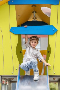 Genç çocuk renkli bir oyun bahçesinde kaydırağın tepesinde diz çöker, korkulukları kavrar ve kendinden emin bir şekilde gülümser..
