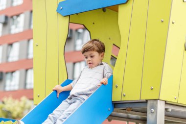 Neşeli küçük çocuk canlı mavi bir kaydırağın üzerinde, apartmanlarla çevrili, modern bir şehir parkında geziyor..