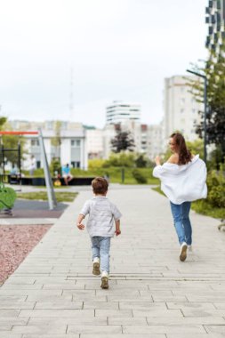 Genç bir çocuk ve annesi, oyun alanları ve binalarla çevrili bir şehir parkında, kaldırımlı bir yolda neşeyle birlikte koşuyorlar..