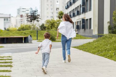 Yeşil kentsel bir mahallede, apartmanları olan bir kaldırımda neşeyle koşan bir anne ve çocuğun arka plan görüntüsü..
