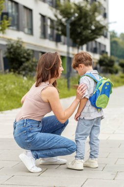 Endişeli anne, üzgün oğluyla konuşmak için diz çöker. Sırt çantası takar ve apartmanların yanındaki kaldırımda destek önerir..