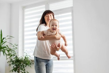 Neşeli anne, neşeli bir şekilde gülen bebeğini bir uçak gibi kaldırır. Eğlencenin ve mutluluğun doğal ışığının tadını çıkarır..